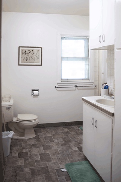 A clean, white bathroom with a toilet, sink, cabinets, shower and a large window with blinds.